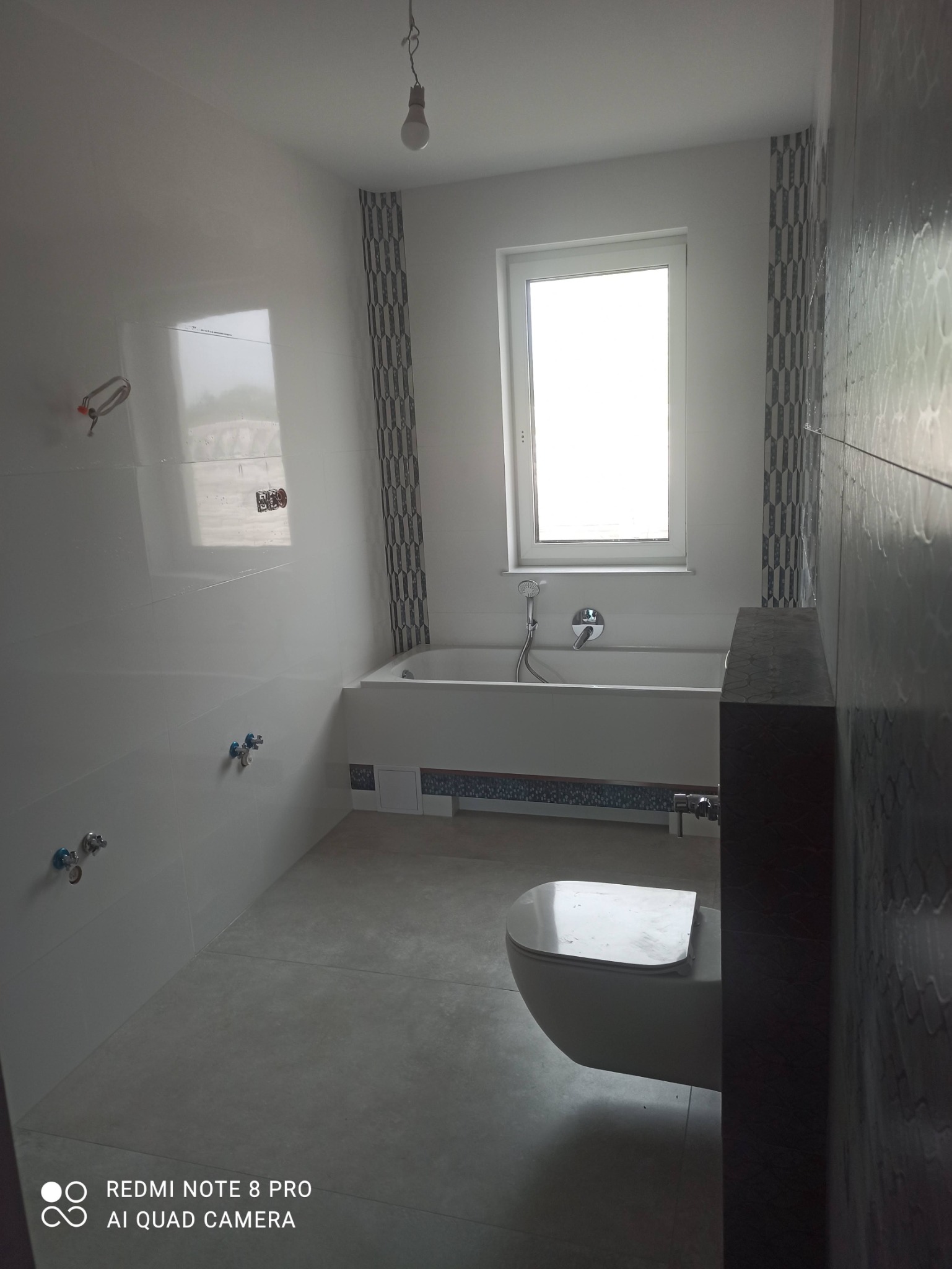 Nowoczesna łazienka z wanną, toaletą i biało-szarą glazurą, widok na okno i surową żarówkę.