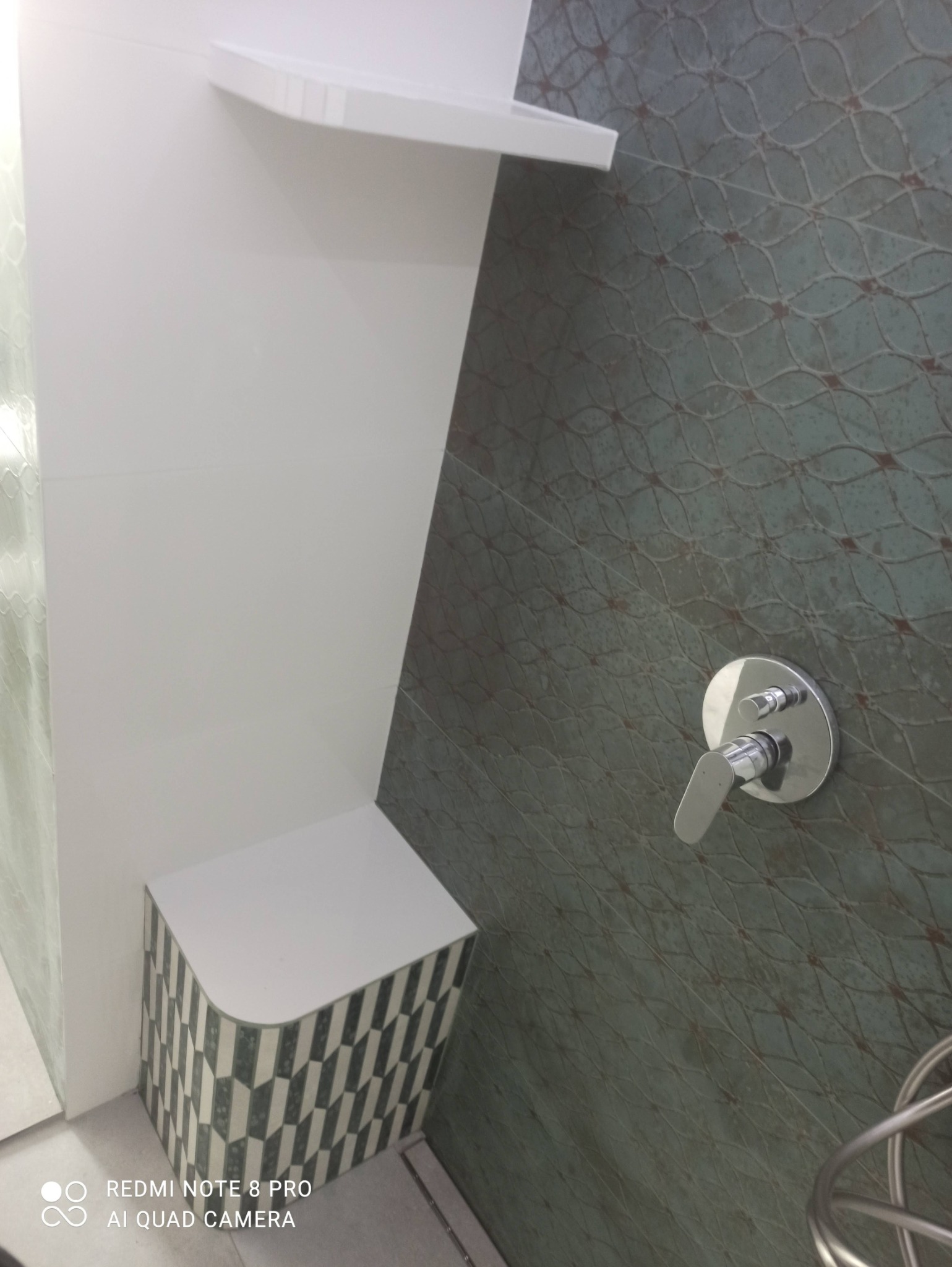 Nowoczesna łazienka z zielono-szarą mozaiką na ścianach, chromowaną armaturą, białą półką i siedziskiem z geometrycznym wzorem.