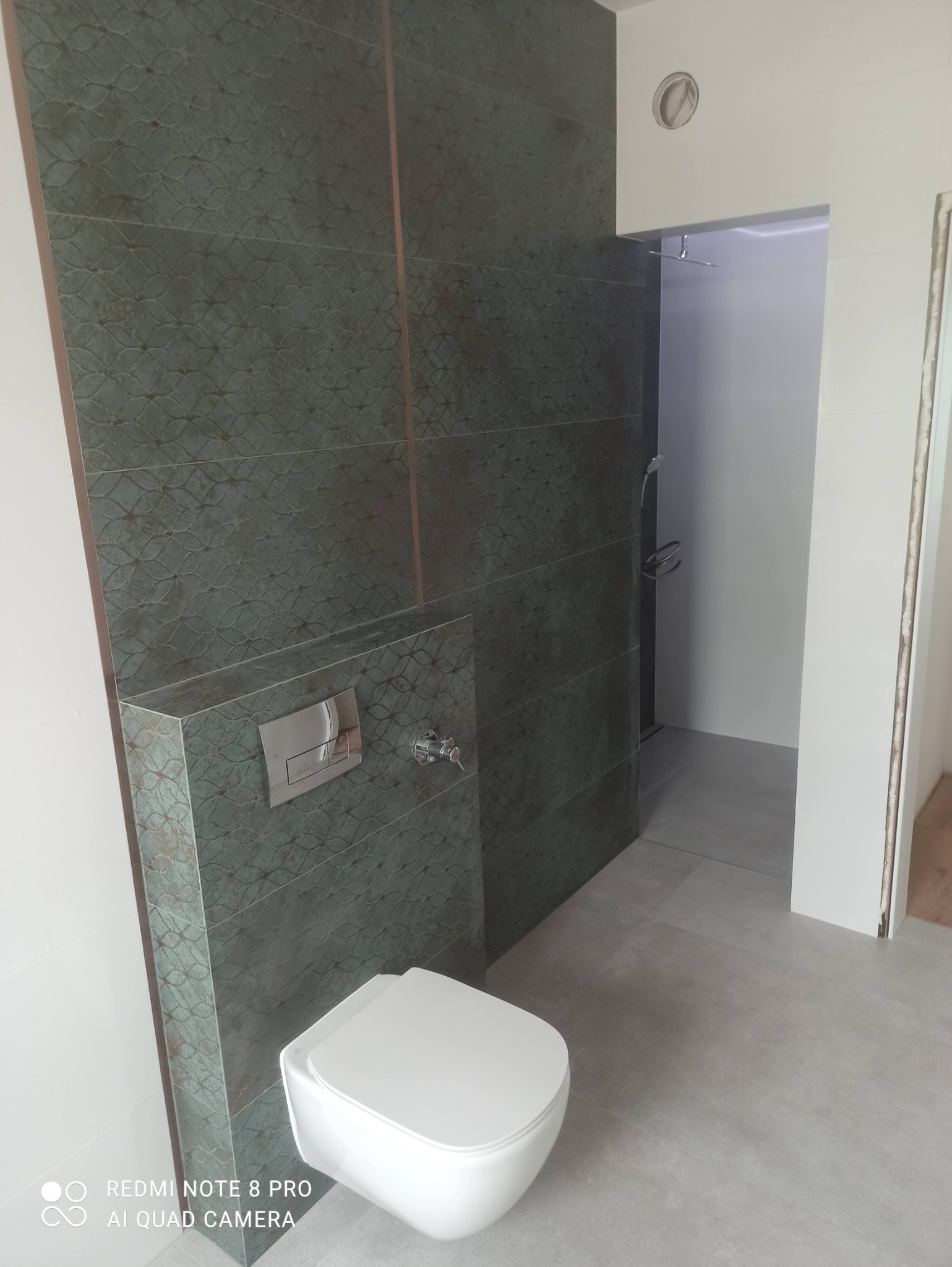 Zawieszona miska WC na tle ściany wyłożonej ciemnozielonymi płytkami z delikatnym wzorem, widoczny chromowany przycisk spłuczki i bateria.