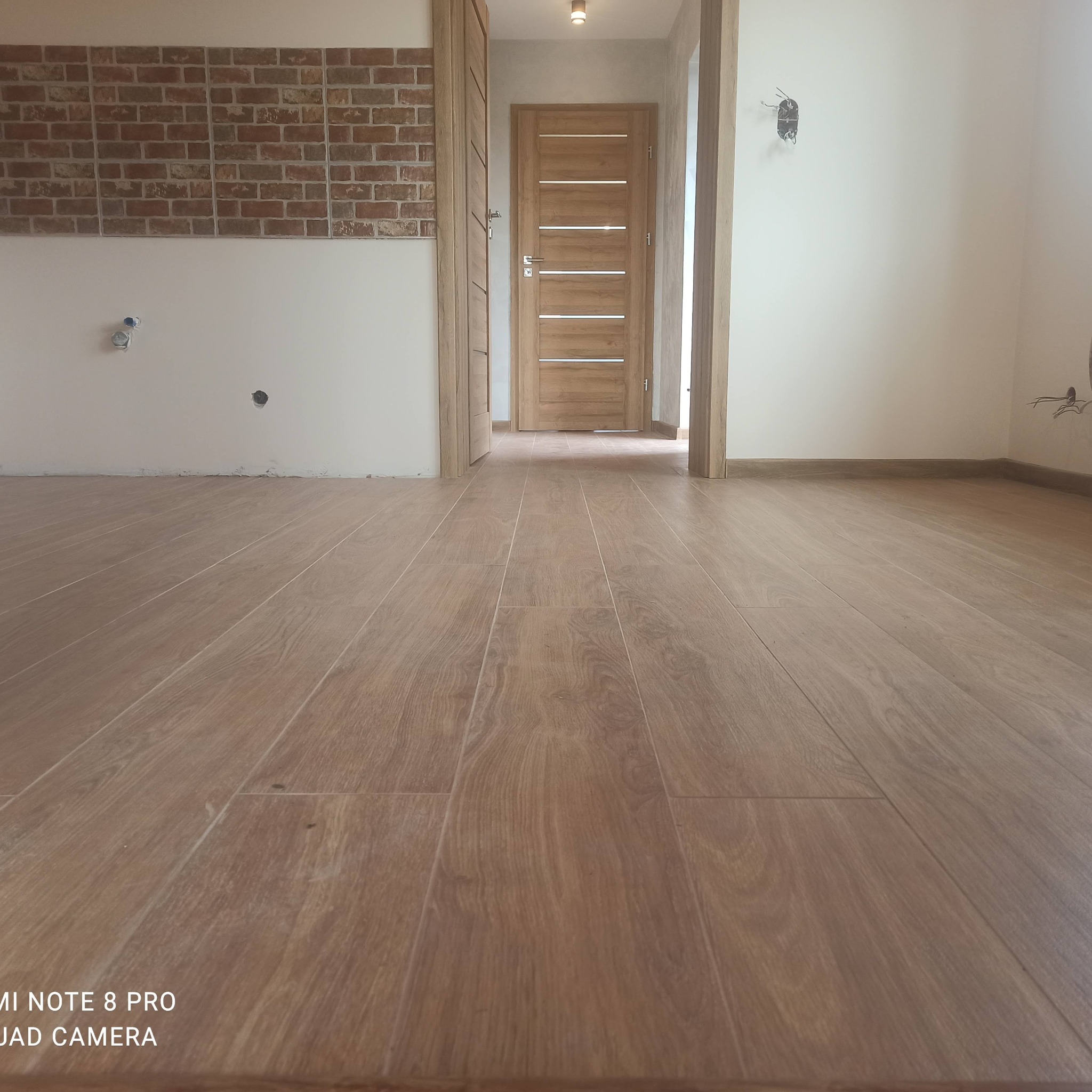 Ułożone panele podłogowe w odcieniach brązu, widoczny fragment ściany z imitacją cegły, drzwi wewnętrzne z poziomymi przeszkleniami i wystającymi kablami elektrycznymi na ścianie.
