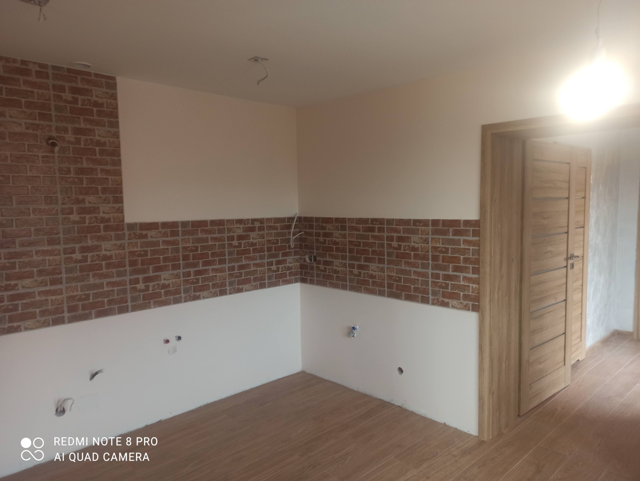 Świeżo wykończone wnętrze z częściowo położoną dekoracyjną czerwoną cegłą na ścianach, widoczne wyprowadzenia elektryczne i hydrauliczne, drewniana podłoga oraz drzwi wewnętrzne z poziomymi frezami.