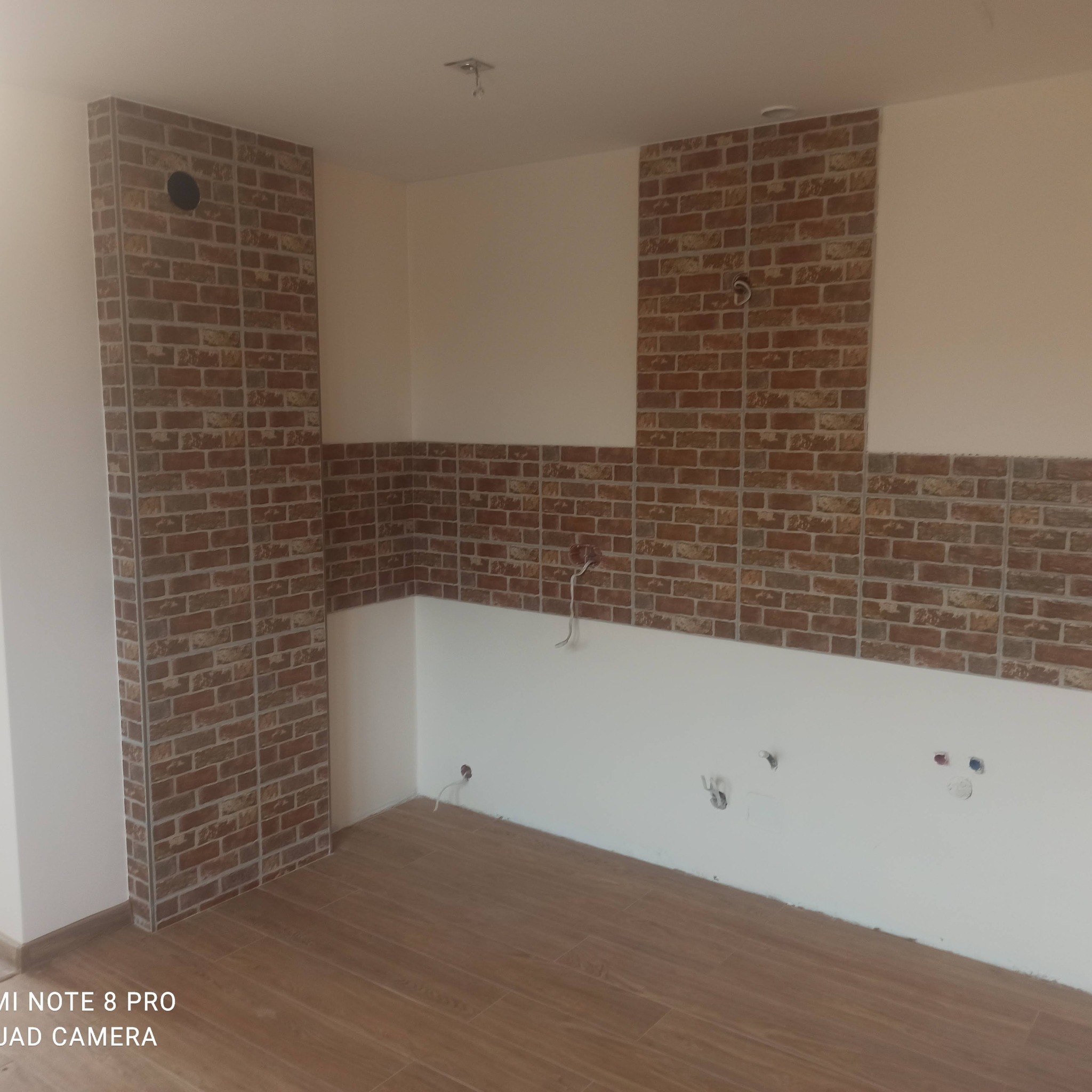 Fragment kuchni w trakcie prac wykończeniowych: ściany częściowo pokryte białą farbą i ceglastymi płytkami, widoczne wyprowadzenia instalacji elektrycznej i wodnej, podłoga wyłożona panelami...