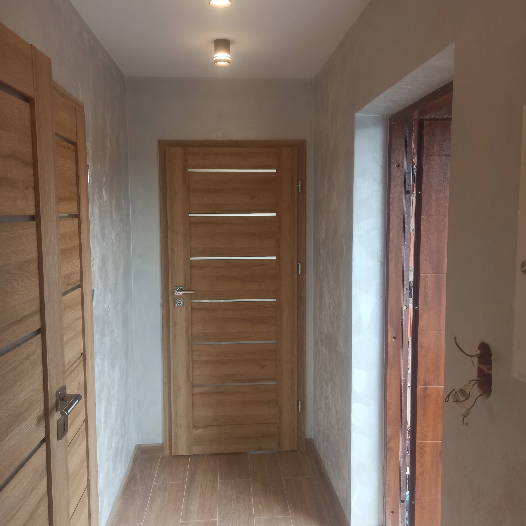 Korytarz po remoncie z trzema drewnianymi drzwiami, płytkami imitującymi drewno na podłodze i dekoracyjną szarą strukturą na ścianach, widoczne niedokończone podłączenie elektryczne przy futrynie.