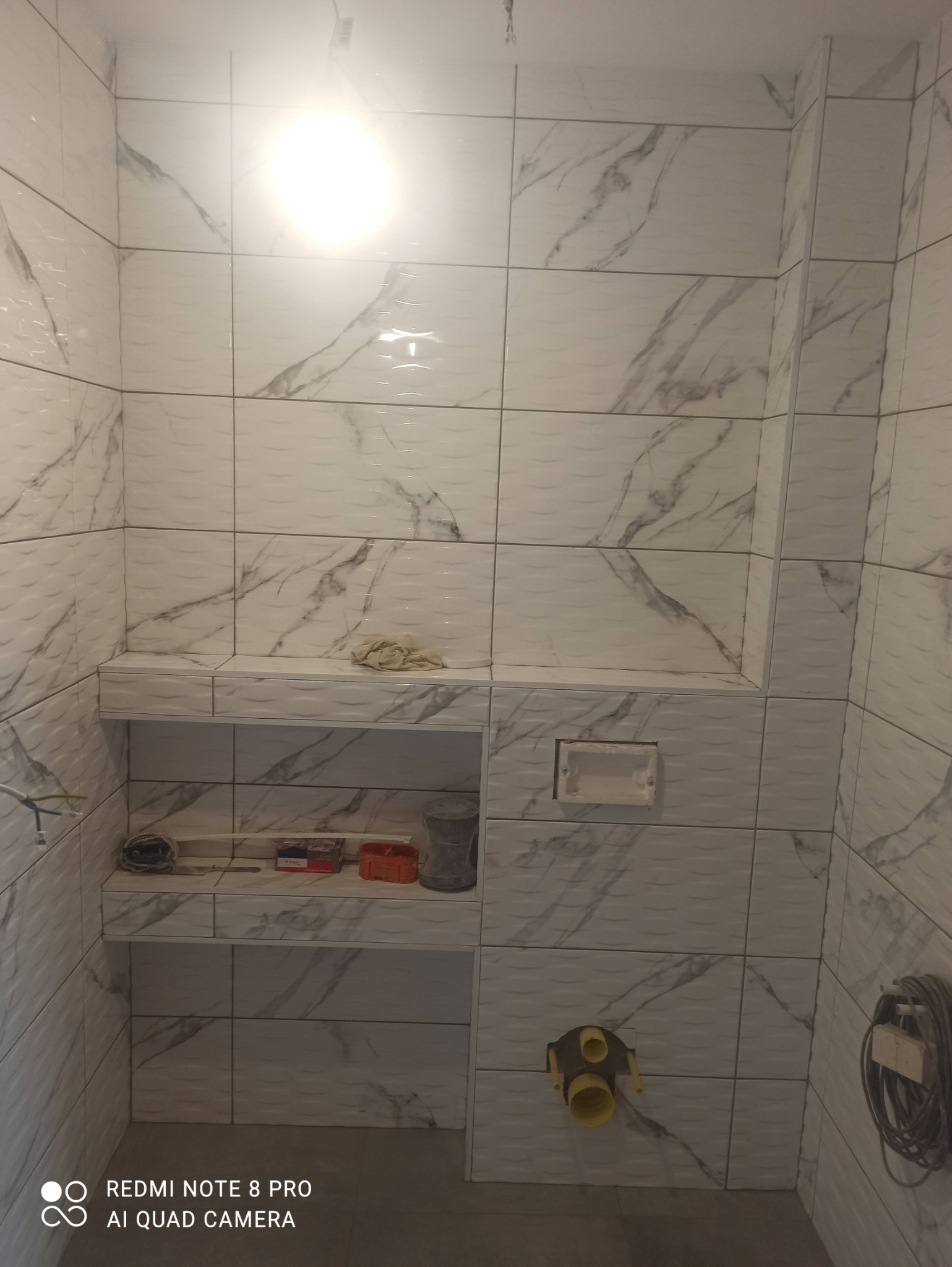 Wykończona ściana łazienki z białymi płytkami imitującymi marmur, z wnęką zabudowaną płytkami, na której leżą akcesoria i narzędzia glazurnicze, widoczny fragment instalacji wodnej.