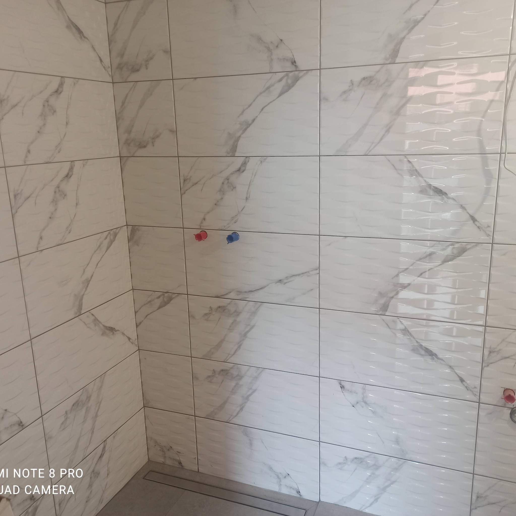 Wykończona ściana łazienki z białych płytek imitujących marmur, z widocznymi przyłączami wodnymi i odpływem liniowym w podłodze.