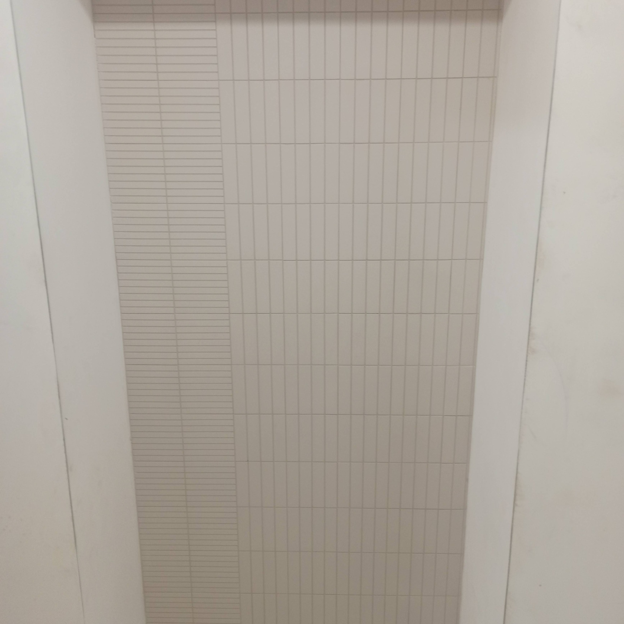 Ściana wykończona jasnymi, pionowo ułożonymi płytkami w kształcie cegiełek, z wąskim pasem płytek poziomych po lewej stronie, otoczona białymi ścianami.