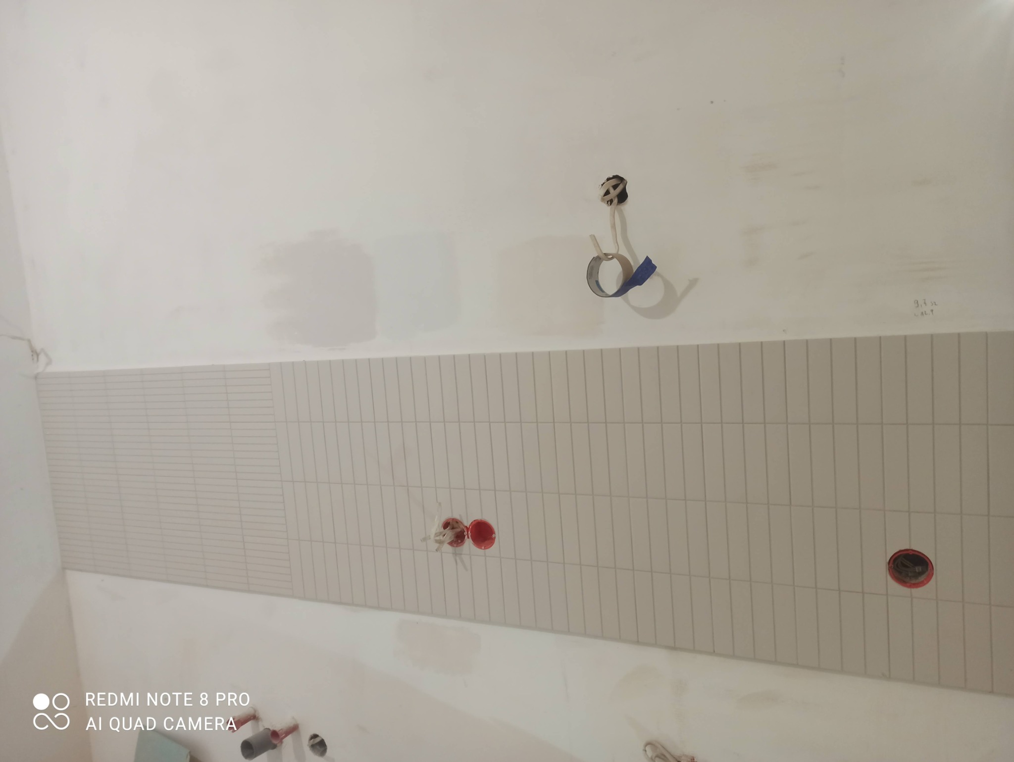 Fragment ściany z ułożonymi białymi, wąskimi płytkami w układzie poziomym, widoczne puszki elektryczne w kolorze czerwonym oraz wystające kable z otworu w ścianie, zabezpieczone taśmą.