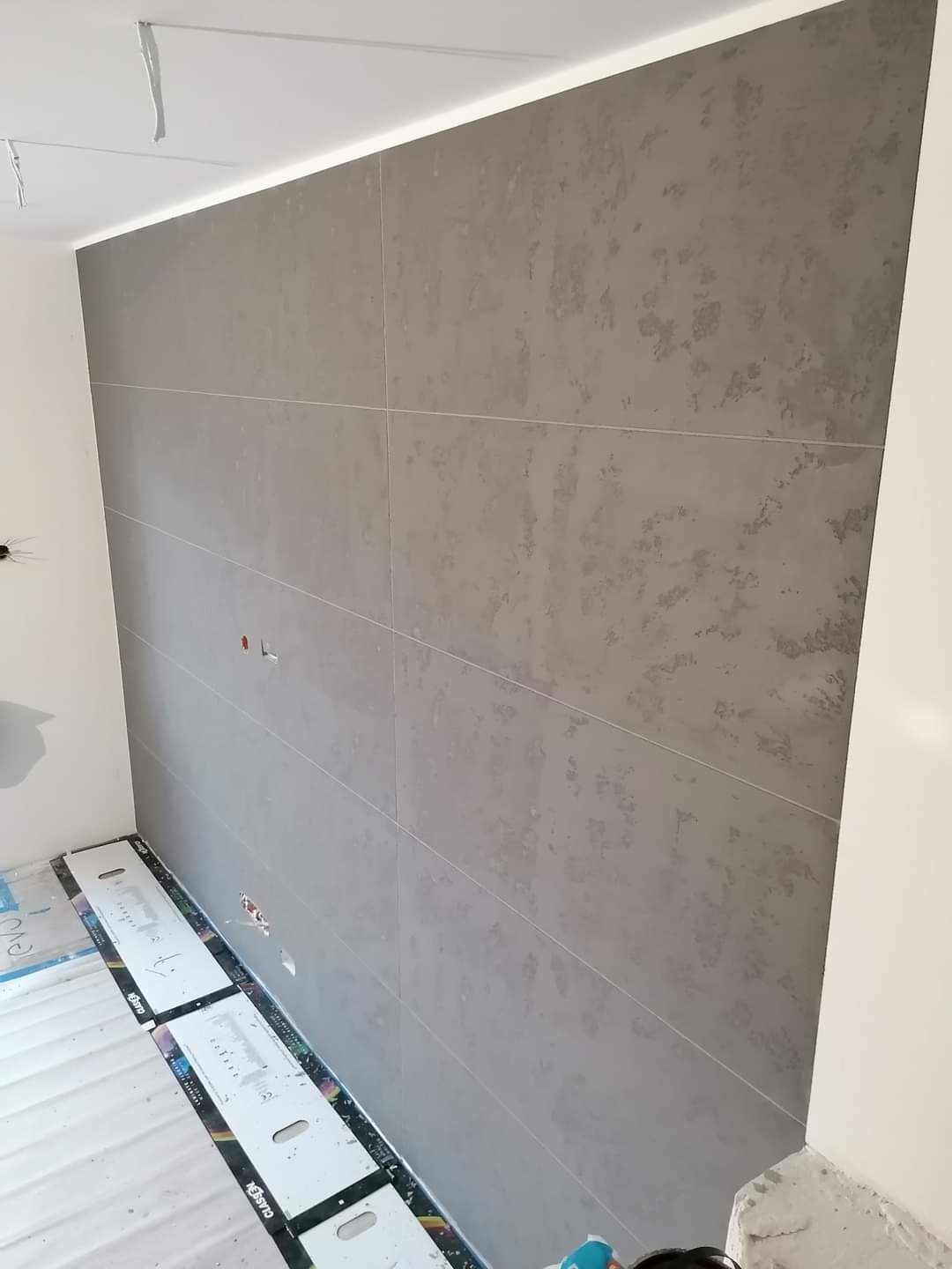 Ściana wyłożona szarymi płytami wielkoformatowymi z delikatnym wzorem, widoczne listwy przypodłogowe i oświetlenie LED podsufitowe, zabezpieczenie podłogi listwami ochronnymi.