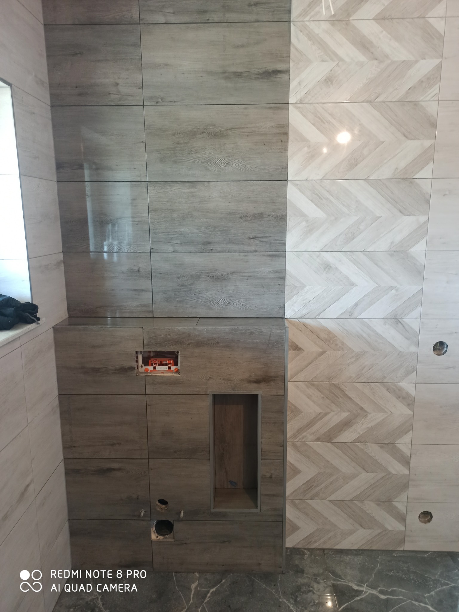 Ściana w łazience wyłożona płytkami imitującymi drewno w dwóch odcieniach: szarym i beżowym, ułożonymi w różne wzory, z widocznymi otworami na instalacje i wnęką w zabudowie.