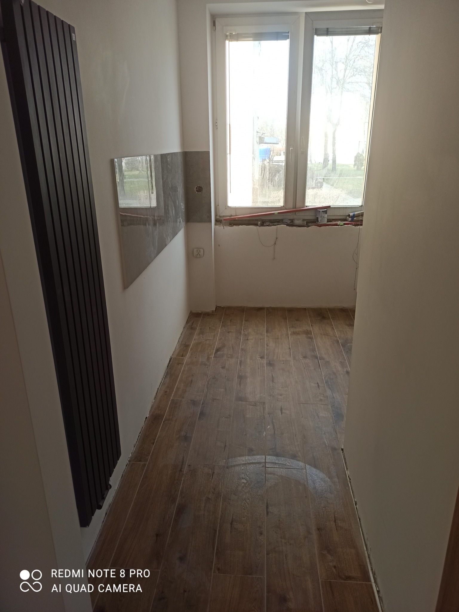 Wnętrze łazienki w trakcie wykańczania: ułożona podłoga z paneli imitujących drewno, częściowo położone płytki na ścianie, grzejnik drabinkowy zamontowany na ścianie, okno z widokiem na zewnątrz...