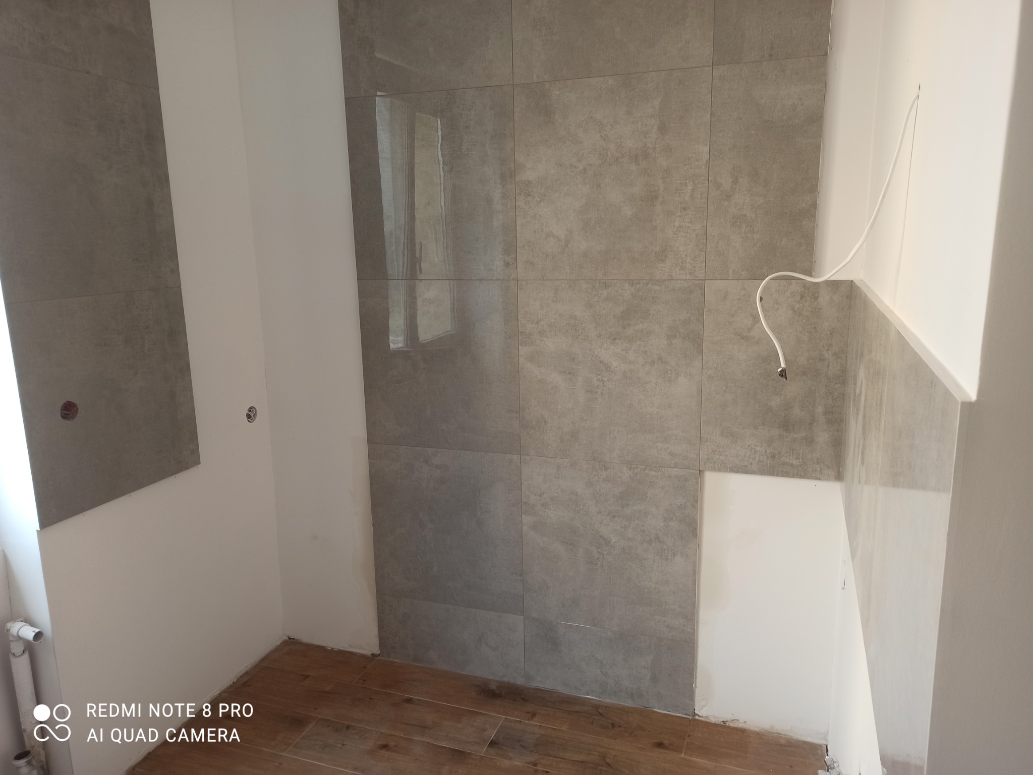 Szare, matowe płytki ścienne w trakcie montażu w łazience, widoczne niedokończone fugi, wystający przewód elektryczny i drewnopodobna podłoga.