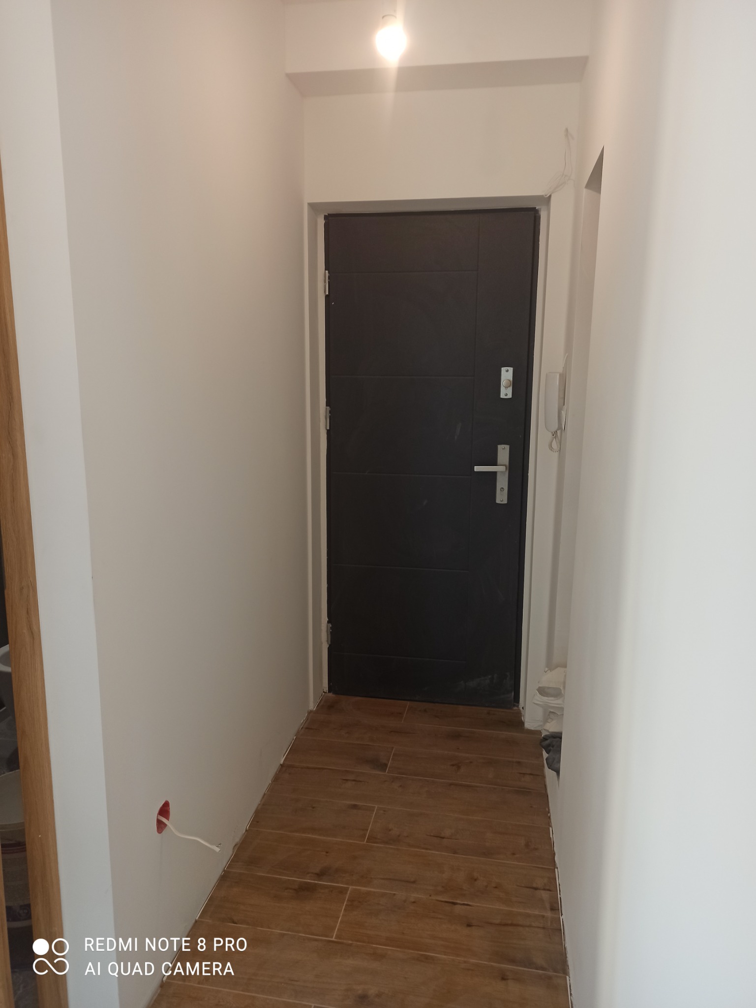 Wnętrze korytarza z ciemnymi drzwiami wejściowymi, brązowymi płytkami podłogowymi imitującymi drewno oraz świeżo pomalowanymi białymi ścianami z widocznym gniazdkiem elektrycznym bez osprzętu.