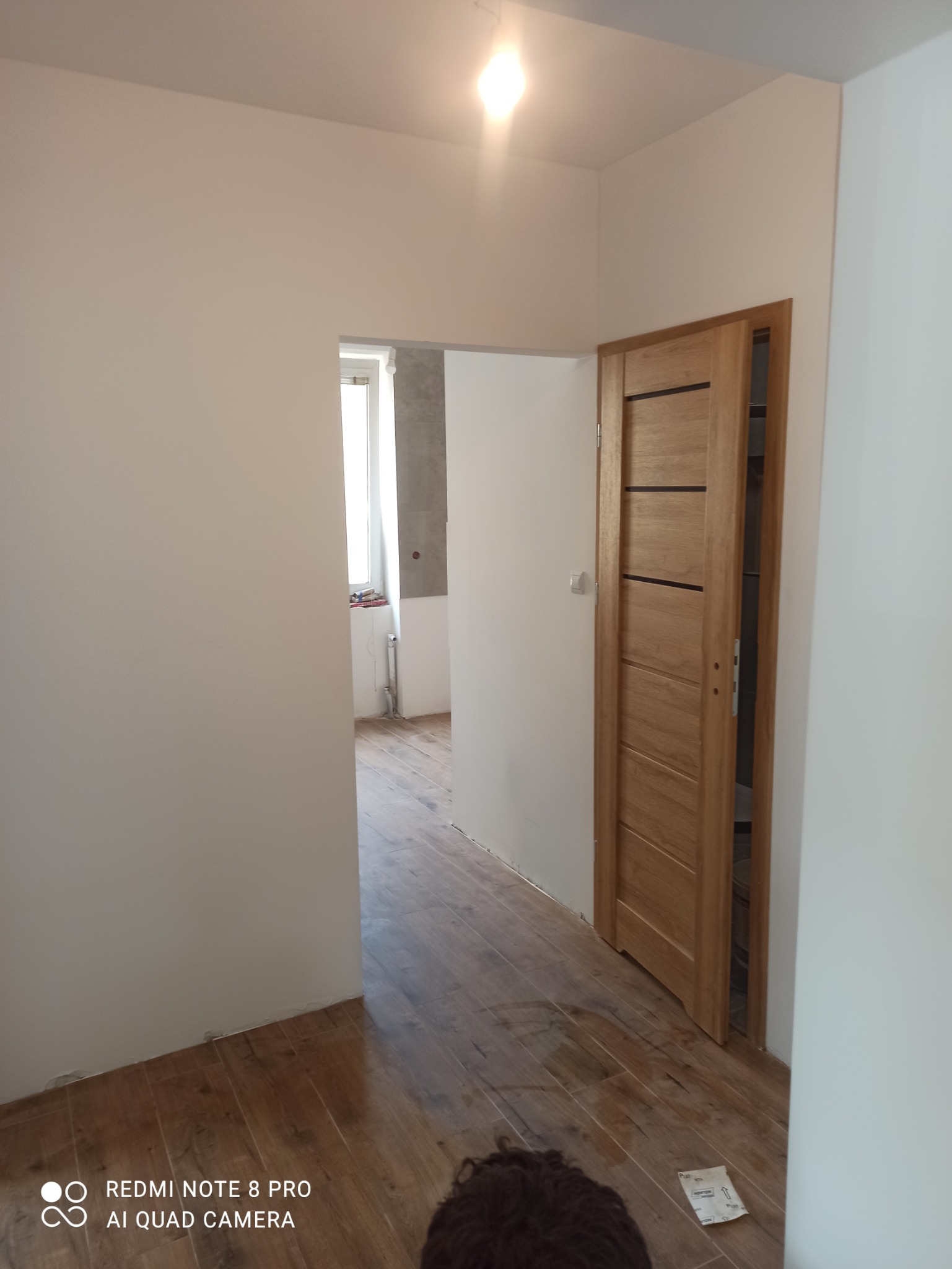 Jasne wnętrze po remoncie: świeżo pomalowane ściany, drewniana podłoga i nowo zamontowane drzwi wewnętrzne z poziomymi wstawkami, widok na wykończoną łazienkę z szarymi płytkami.