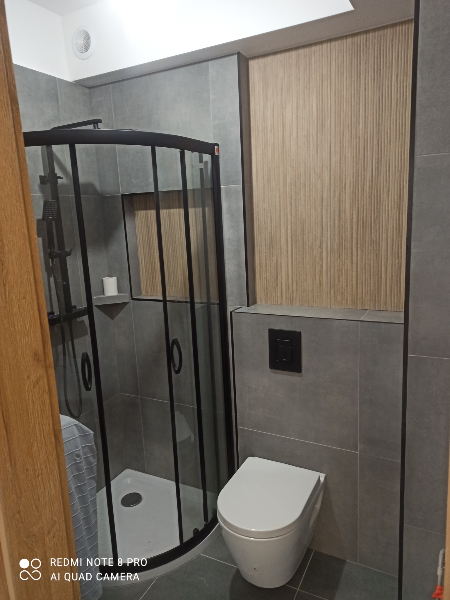 Nowoczesna łazienka z kabiną prysznicową z czarnymi profilami, wiszącą toaletą i szarymi płytkami, akcentowana drewnopodobną okładziną ścienną.