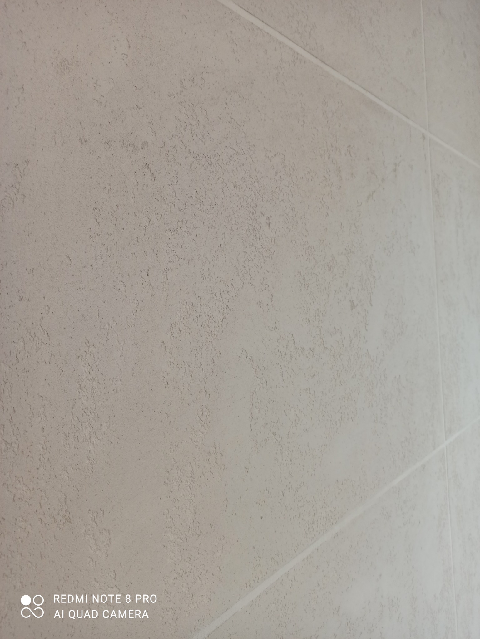 Ściana wykończona jasnobeżową, strukturalną farbą dekoracyjną z widocznymi fugami między płytami.