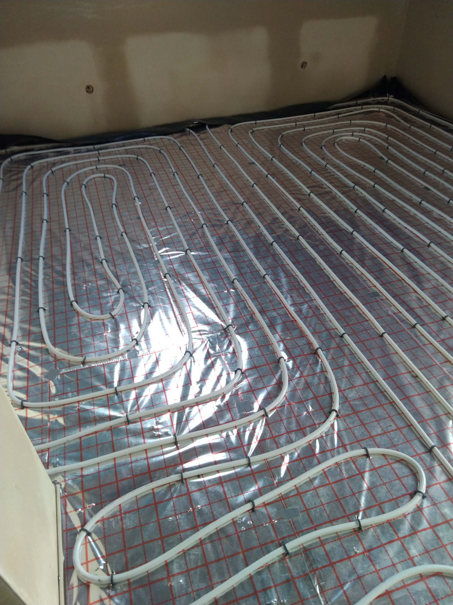 Białe rury ogrzewania podłogowego ułożone na srebrnej folii z czerwoną siatką, zamocowane czarnymi klipsami, widok z góry w surowym wnętrzu.