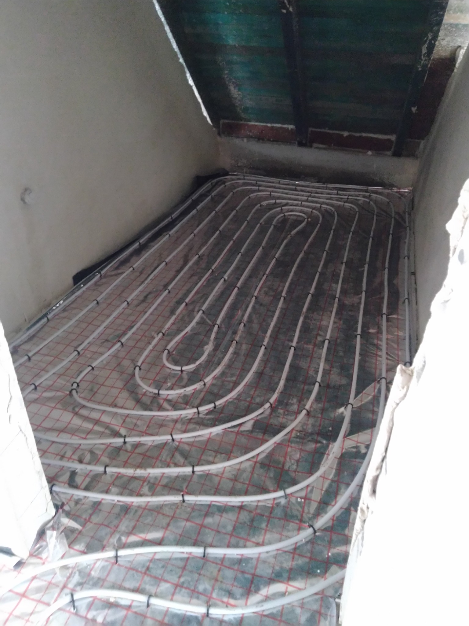 Białe rury ogrzewania podłogowego ułożone w pętle na folii z czerwoną siatką, widok z góry na surowy stan wykończenia wnętrza.