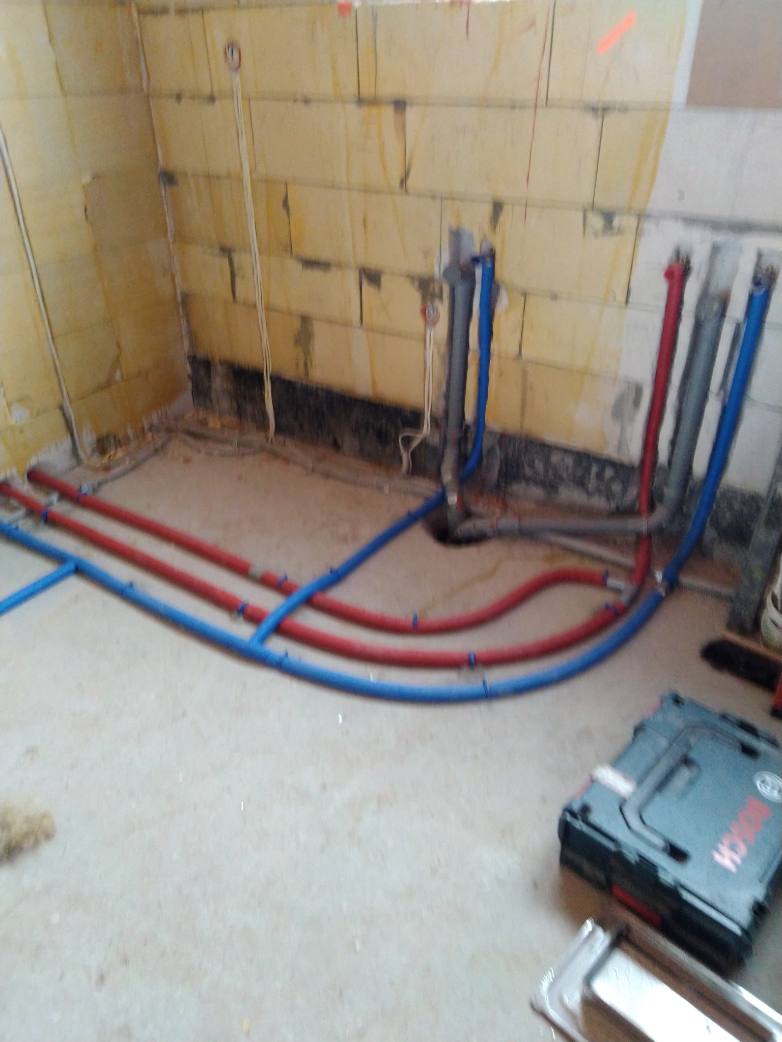 Wnętrze w trakcie prac hydraulicznych: widoczne rury z tworzywa w kolorze niebieskim i czerwonym, wychodzące ze ściany z pustaków, skrzynka narzędziowa Bosch w tle.