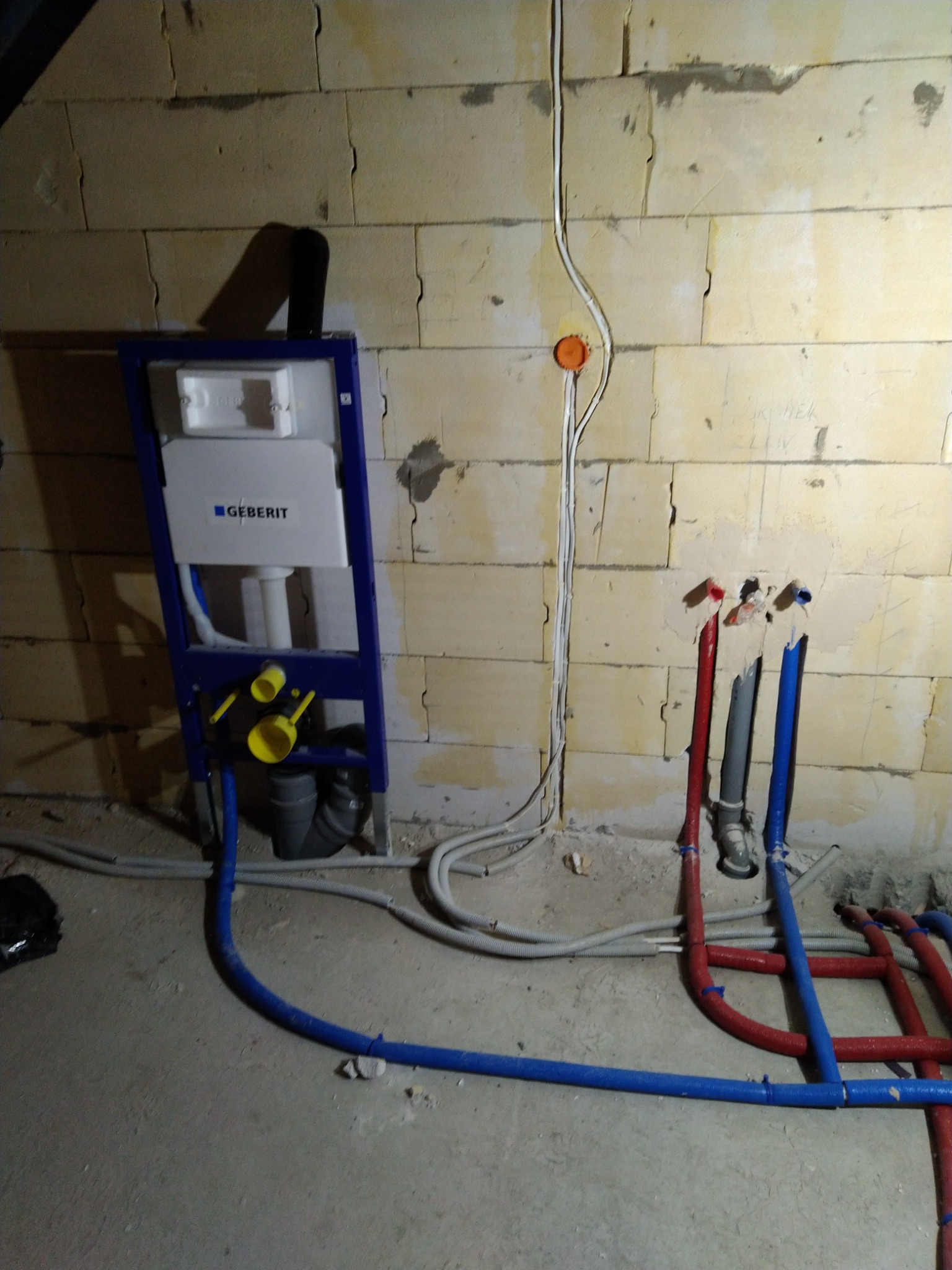Stelaż podtynkowy Geberit zamontowany przy surowej ścianie z betonu komórkowego, widoczne rury instalacji wodnej w kolorach czerwonym i niebieskim oraz instalacja elektryczna w peszlu.