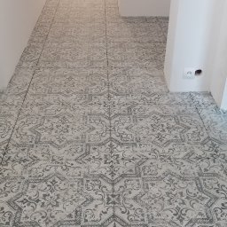 PANORAMA MARCIN KWITOWSKI - Ułożona podłoga z płytek ceramicznych w szaro-białe, dekoracyjne wzory w korytarzu, widoczny grzejnik zabezpieczony folią malarską w tle.