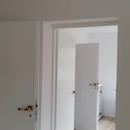 Wnętrze mieszkania w Łodzi z dwiema białymi, częściowo otwartymi drzwiami z złotymi klamkami i panelami podłogowymi imitującymi jasne drewno.