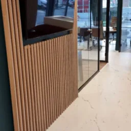 Nowoczesne biuro z drewnianą ścianką działową z pionowych listew, na której zawieszony jest telewizor. W tle przeszklona sala konferencyjna z krzesłami i stołem. Podłoga wyłożona płytkami...