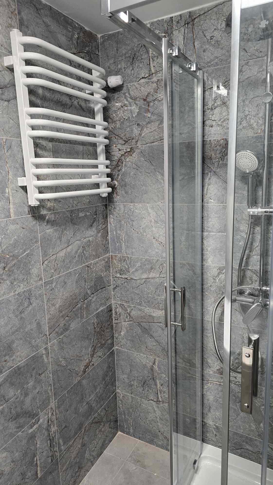 Szara łazienka z marmurowymi płytkami, biały grzejnik drabinkowy i kabina prysznicowa z przezroczystego szkła z chromowanymi okuciami.
