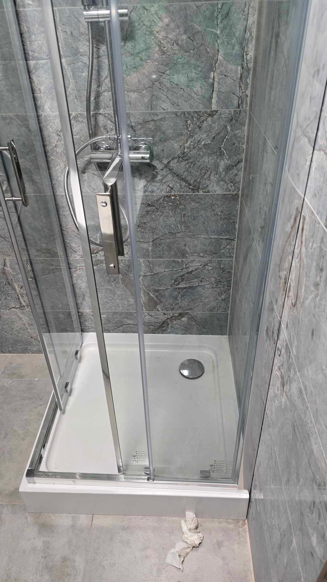 Szklana kabina prysznicowa z chromowanymi detalami na tle szarych, marmurkowych płytek. Widoczny biały brodzik i fragment podłogi z resztkami po montażu.