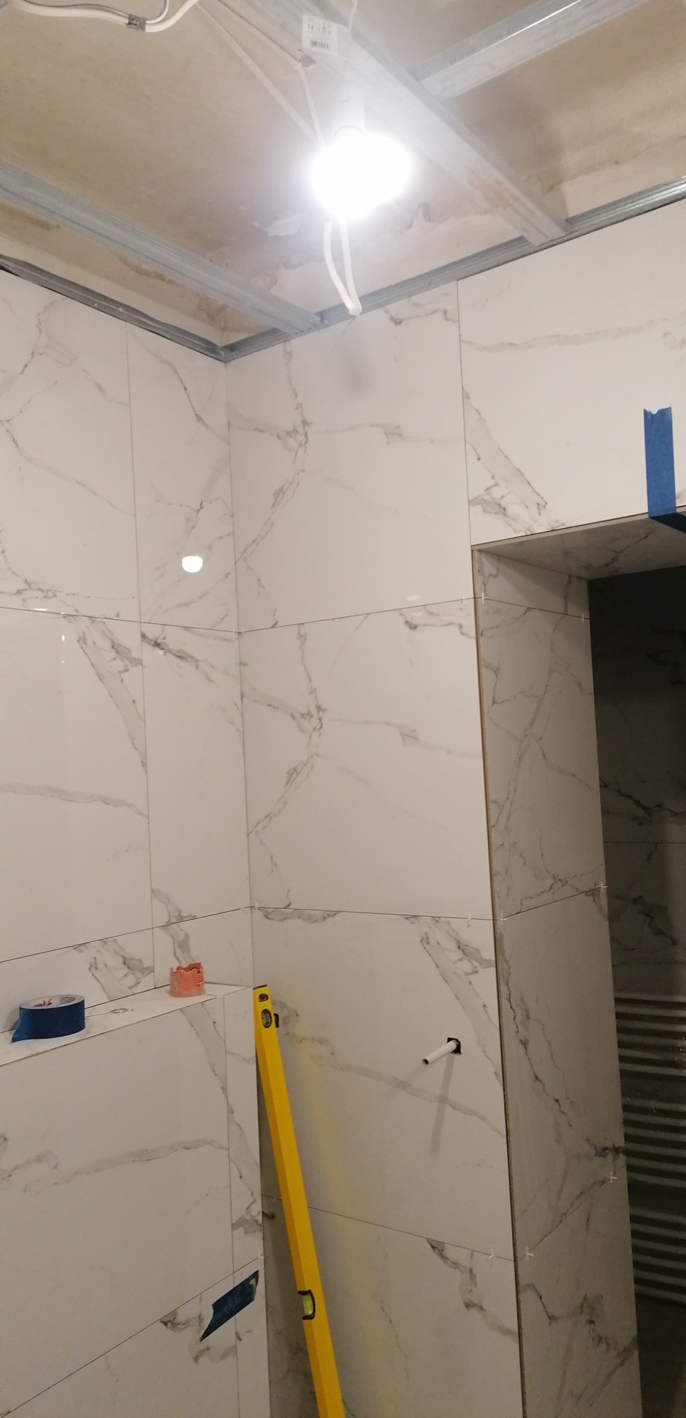 Układanie białych płytek imitujących marmur w łazience, widoczna poziomica, taśma malarska i fragment instalacji elektrycznej pod sufitem.