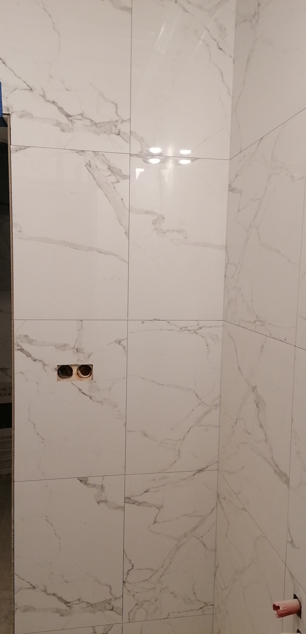 Ściana w trakcie układania płytek imitujących marmur, widoczne otwory na gniazdka elektryczne i fragment rury w dolnym rogu.