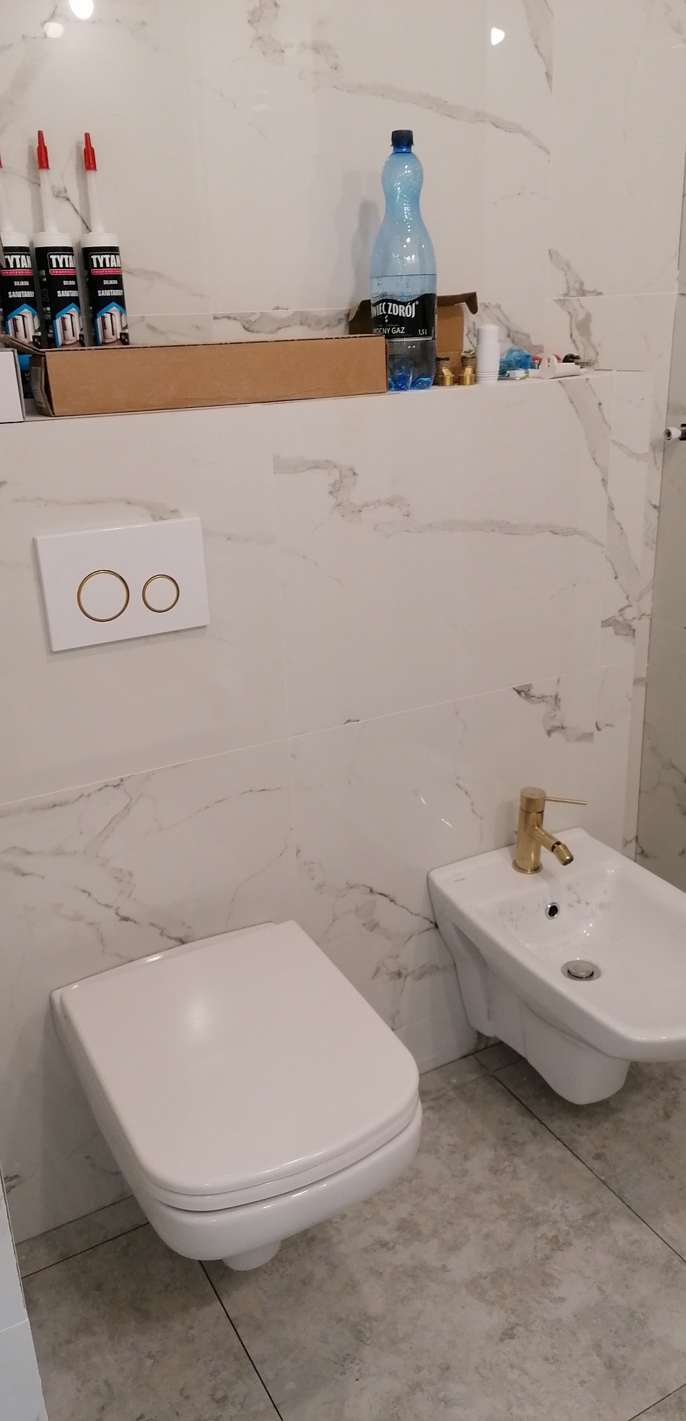 Nowa, jasna łazienka z marmurowymi płytkami, wiszącą toaletą, bidetem z złotą armaturą i przyciskiem spłukiwania w kolorze białym i złotym. Na półce nad toaletą widoczne materiały budowlane...