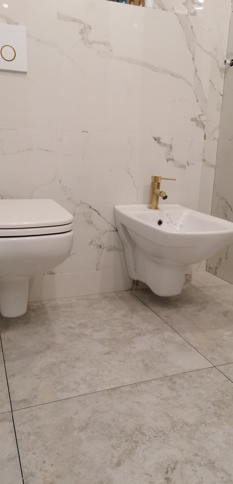 Bidet z złotą armaturą obok toalety na tle ściany wyłożonej białymi płytkami imitującymi marmur, widok z dołu.