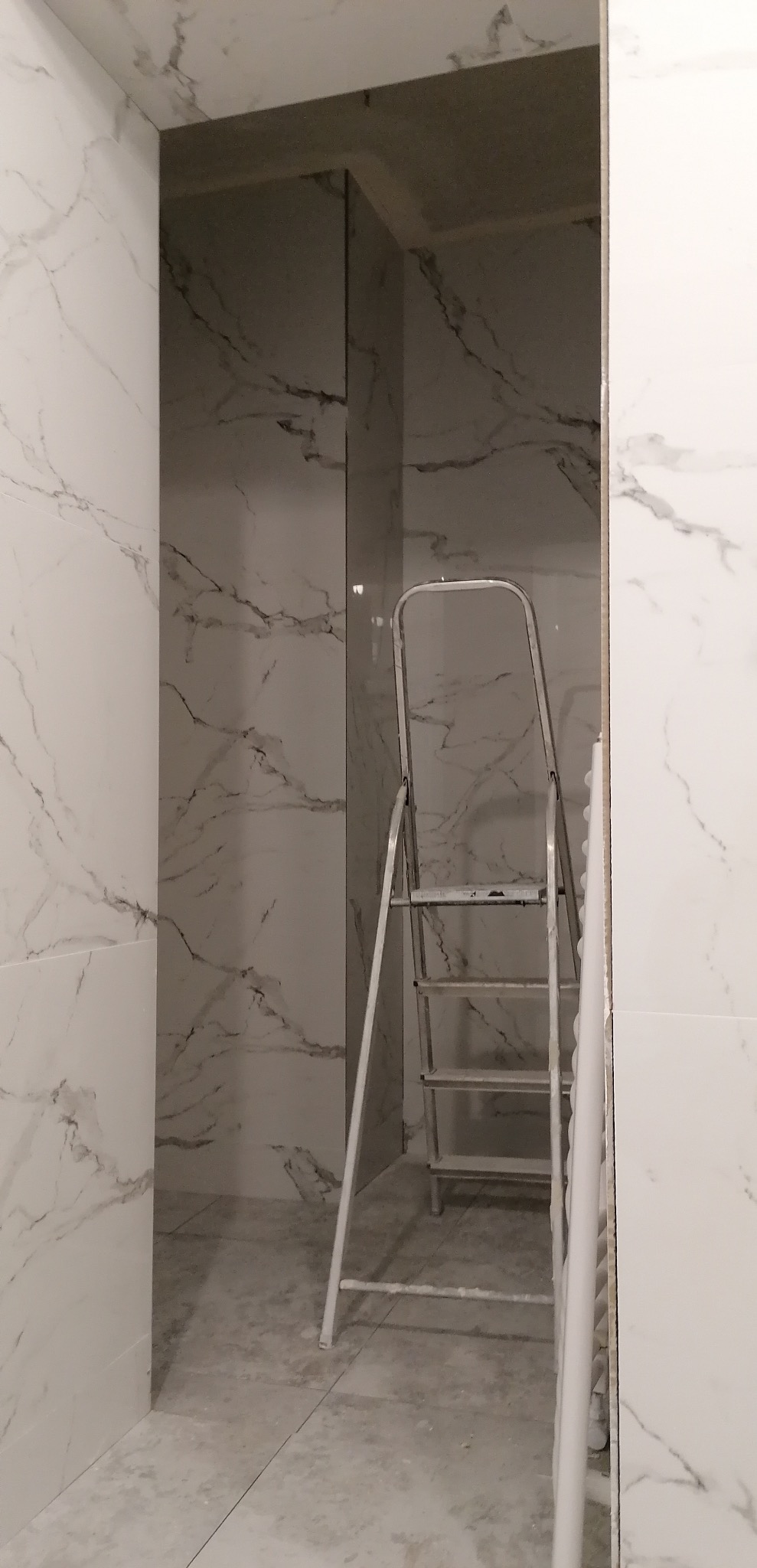 Wnętrze łazienki w trakcie prac wykończeniowych, widoczne białe płytki imitujące marmur na ścianach, aluminiowa drabina oparta o ścianę oraz fragment listwy przypodłogowej w rogu.