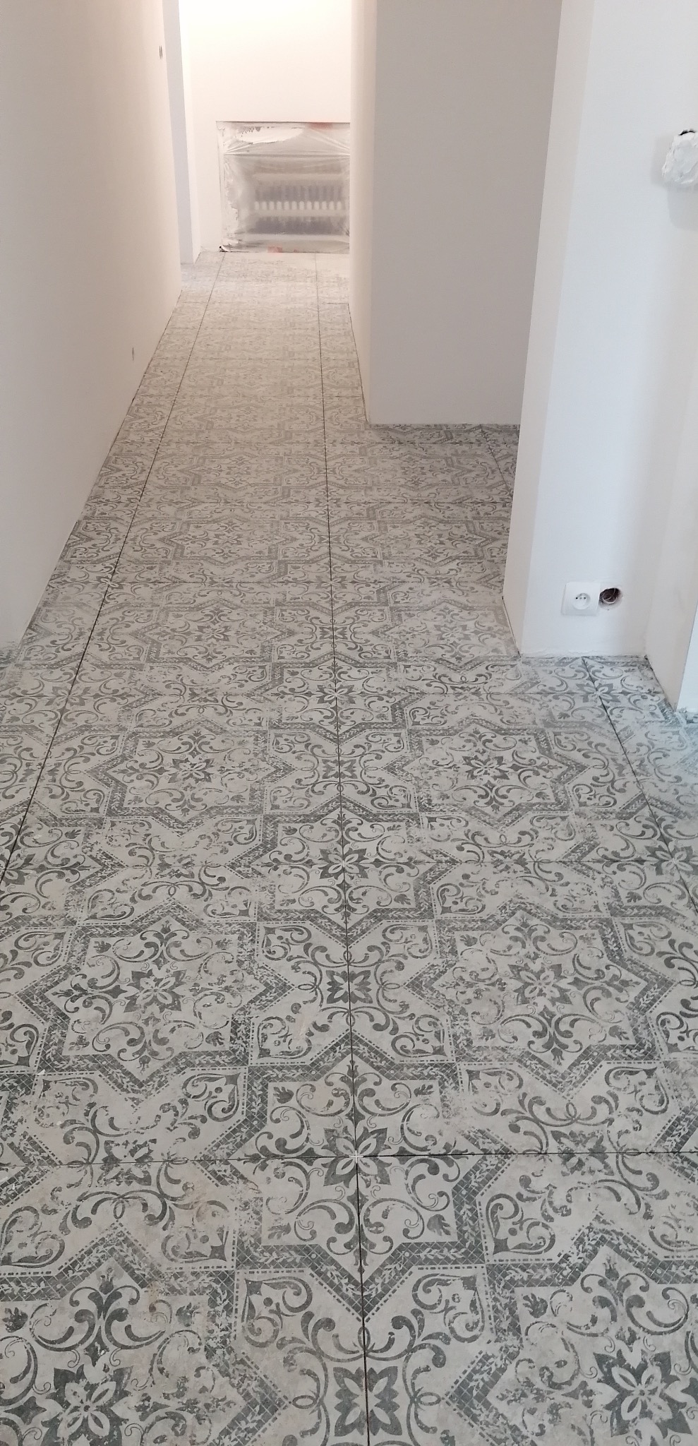 Ułożona podłoga z płytek ceramicznych w szaro-białe, dekoracyjne wzory w korytarzu, widoczny grzejnik zabezpieczony folią malarską w tle.