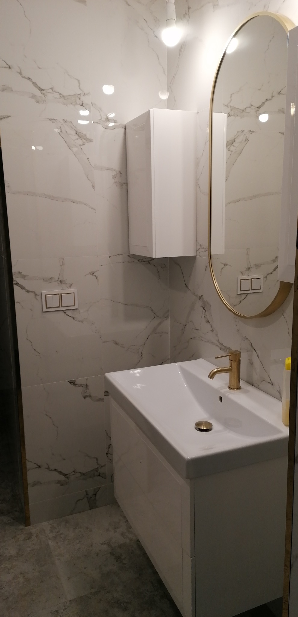 Nowoczesna łazienka z białą szafką pod umywalką, złotą armaturą i owalnym lustrem w złotej ramie na tle marmurowych płytek.