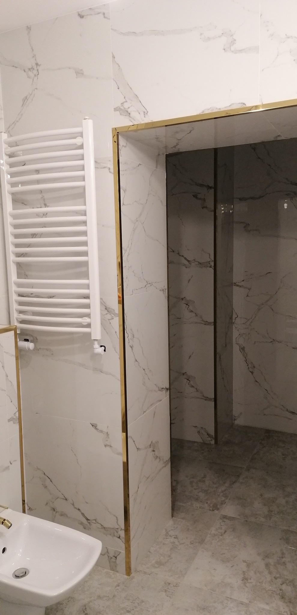 Wykończona łazienka z białymi marmurkowymi płytkami, złotymi listwami wykończeniowymi, białym grzejnikiem drabinkowym i bidetem.