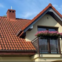 Czerwona dachówka na domu z balkonem, na którym stoją donice z różowymi kwiatami, widoczny komin i okno dachowe.