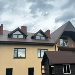 Dom z beżową elewacją i brązowym dachem z blachy trapezowej, widoczne kominy z cegły i okna z czarnymi ramami, na tle pochmurnego nieba.