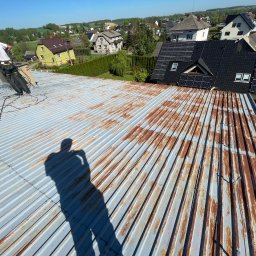 Minor Malowanie Dachów Natalia Szymczak - Pracownik w trakcie mycia zardzewiałego dachu z blachy falistej za pomocą myjki ciśnieniowej. Widoczny cień osoby robiącej zdjęcie, w tle zabudowa jednorodzinna.