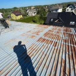 Pracownik w trakcie mycia zardzewiałego dachu z blachy falistej za pomocą myjki ciśnieniowej. Widoczny cień osoby robiącej zdjęcie, w tle zabudowa jednorodzinna.