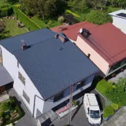 Widok z góry na dom z ciemnoszarym, odnowionym dachem z blachy trapezowej. Obok dom z dachem w kolorze ceglastym. Na podjeździe stoi bus firmy budowlanej.