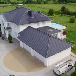 Widok z góry na dom z ciemnoszarym dachem i białymi ścianami. Na dachu panele słoneczne. Obok domu zaparkowany bus z drabiną.