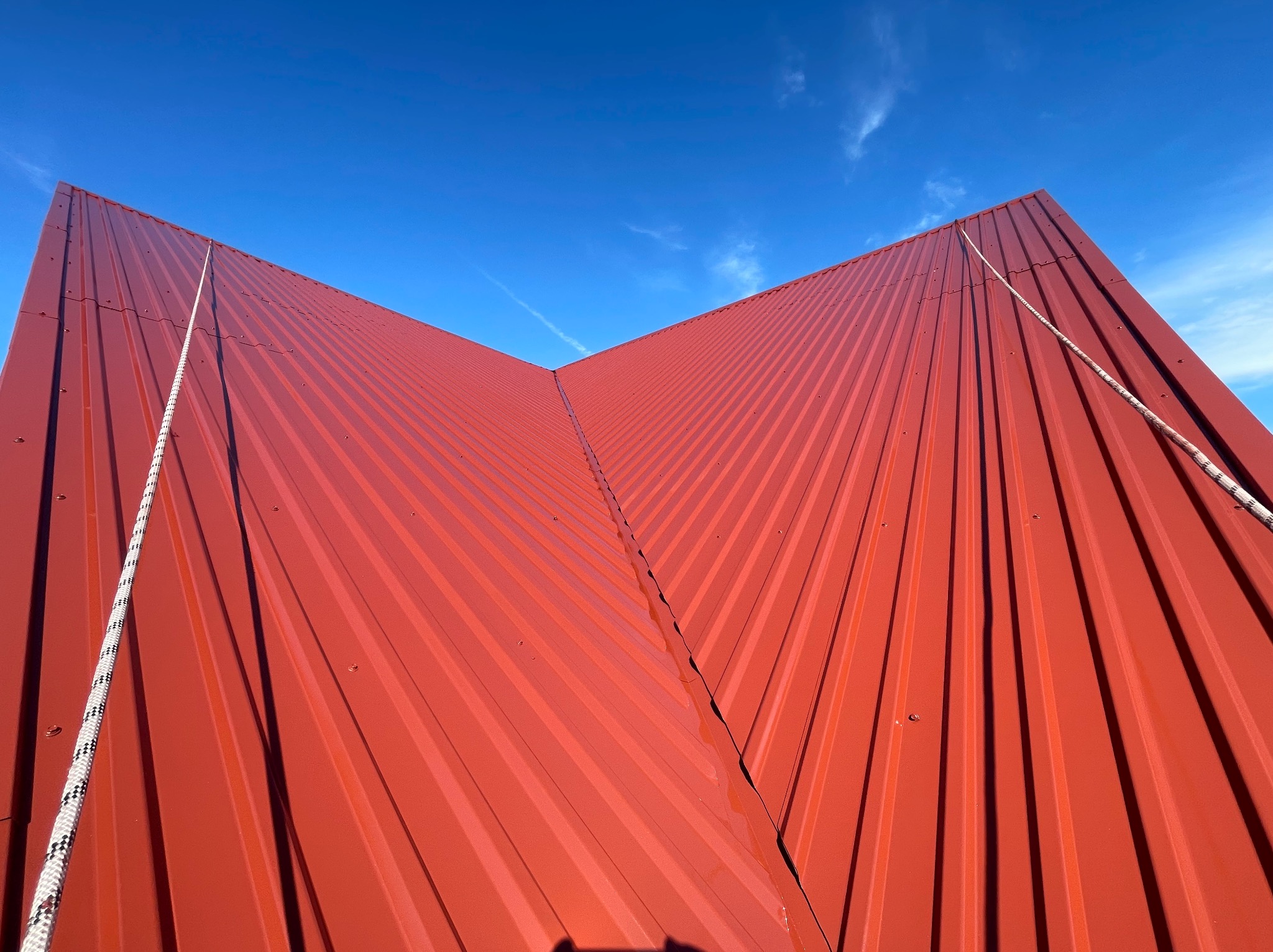 Czerwony, blaszany dach z wyraźnymi żłobieniami pod kątem, liny zabezpieczające zwisają z krawędzi na tle błękitnego nieba.