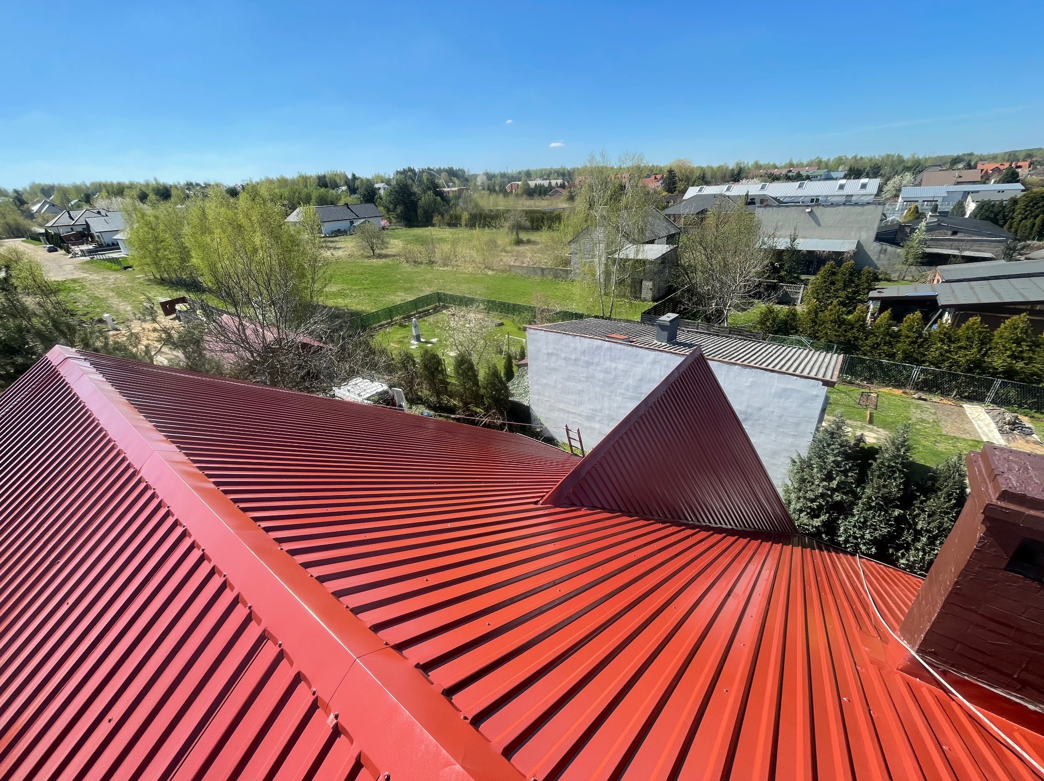 Czerwony, blaszany dach po malowaniu, widok z góry na tle zabudowań i zieleni w słoneczny dzień. Charakterystyczne żłobienia blachy.