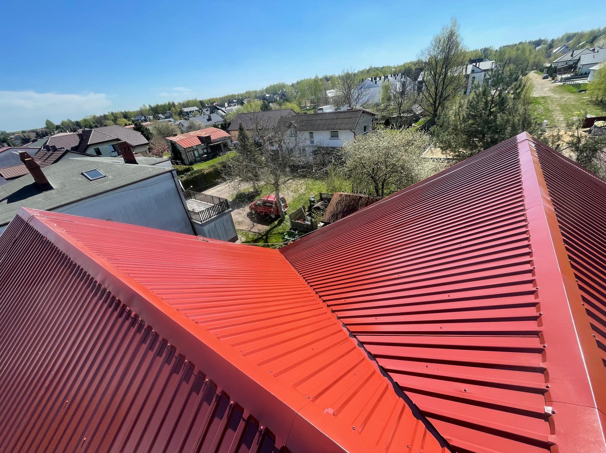 Czerwony, blaszany dach domu w słoneczny dzień. Widok z góry na regularne linie blachy i otoczenie domów oraz zieleni. Dach po renowacji lub nowo pomalowany.