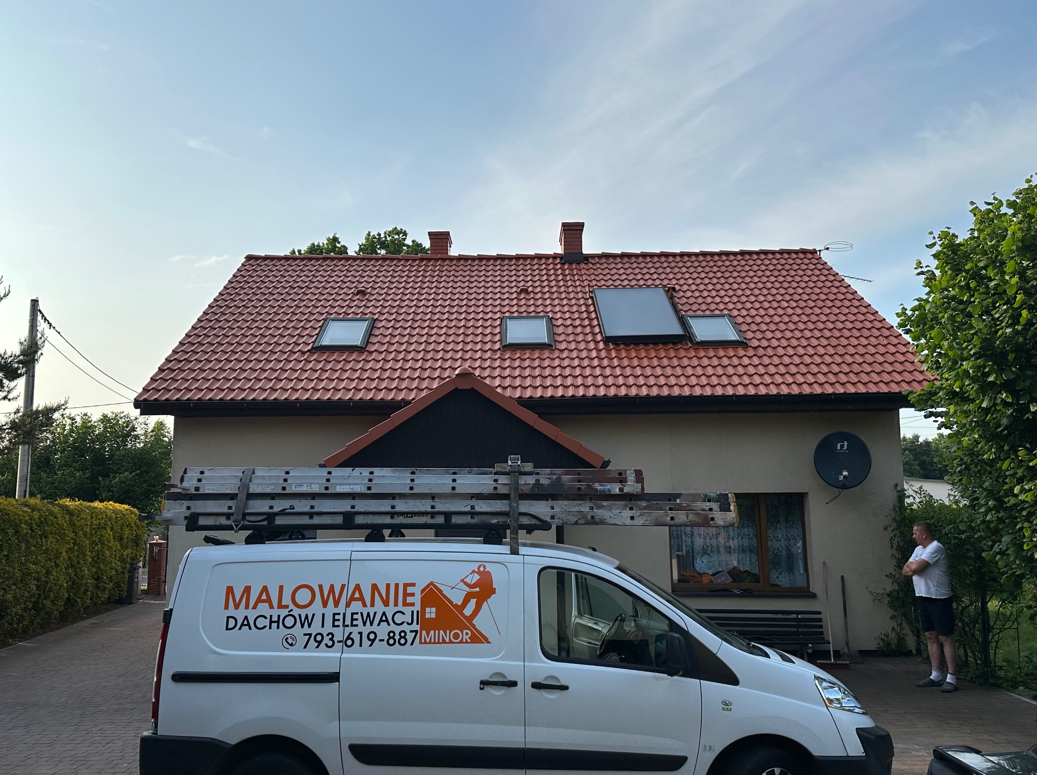 Dom z czerwonym dachem i oknami dachowymi, na tle błękitnego nieba. Na pierwszym planie biały samochód z reklamą firmy remontowej.