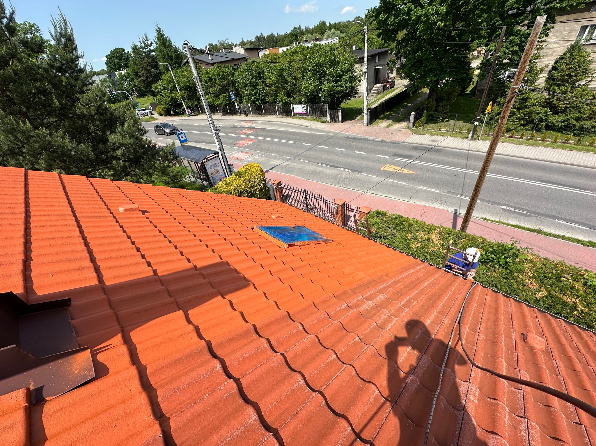 Czerwony dach z dachówki ceramicznej po malowaniu, widok z góry na tle ulicy i zieleni. Widoczny cień osoby i narzędzia pracy.