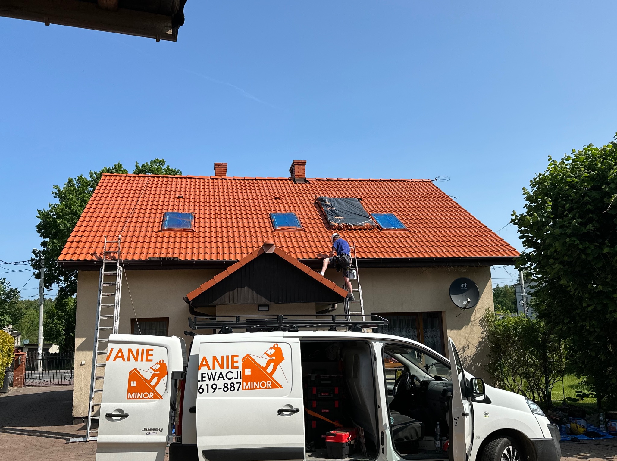 Pracownik na drabinie, maluje fragment dachu domu z czerwoną dachówką. Widoczny samochód firmowy z logo i numerem telefonu. Część dachu zabezpieczona folią.