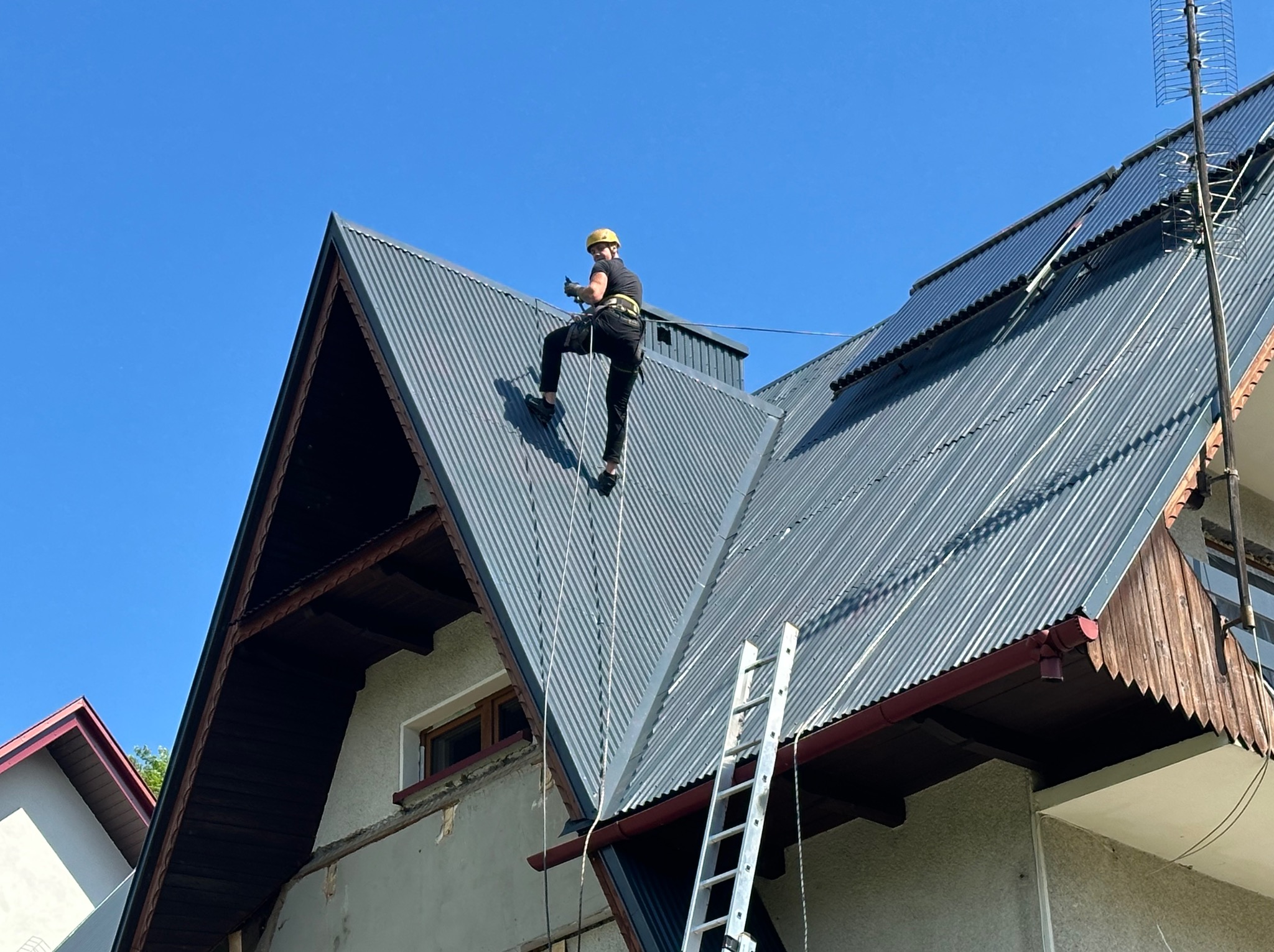 Pracownik w kasku, zabezpieczony linami, na spadzistym dachu z blachy falistej. Widoczna drabina i fragment elewacji budynku. Błękitne niebo w tle.