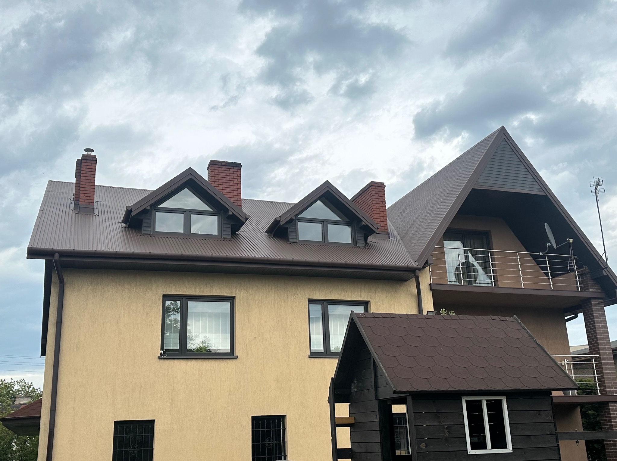 Dom z beżową elewacją i brązowym dachem z blachy trapezowej, widoczne kominy z cegły i okna z czarnymi ramami, na tle pochmurnego nieba.